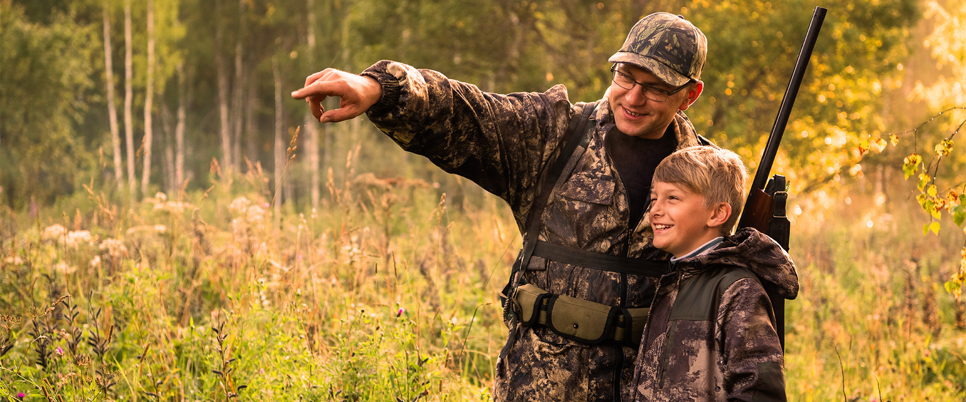 Ein Erwachsener mit Gewehr und ein Junge in Jagdkleidung im Wald, der Erwachsene zeigt auf etwas im Freien, vermittelt Jagdwissen – ideal für das realitätsnahe Jagdtraining von Schiess-Kino Cramberg für Jäger und Jungjäger.